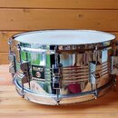 Stříbrný kovový snare drum s chromovaným povrchem. Na boku je černý čtvercový štítek s bílým nápisem MAPEX a menším textem pod ním, který je nečiteln…