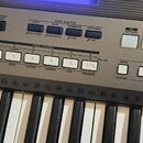Detailní záběr na ovládací panel a klávesy elektronického klavíru v šedé barvě. Na panelu jsou viditelné nápisy jako PORTABLE GRAND, VOICE CONTROL (S…