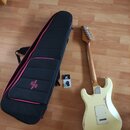 Kremově bílá elektrická kytara s tvarem typu Stratocaster, položená na dřevěné podlaze. Na zadní straně těla jsou viditelné oděrky a opotřebení laku.…