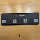 Černý obdélníkový MIDI kontrolér s nápisem iRig BlueBoard. Zařízení má čtyři bílá tlačítka označená písmeny A, B, C a D. V pravém horním rohu je vidě…
