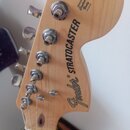 Záběr na hlavu elektrické kytary z přírodního dřeva. Na povrchu je černý nápis Fender® STRATOCASTER a menší text ORIGINAL Contour Body PATENTED. Jsou…