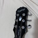Černá hlava kytary s nápisem Epiphone na horní části a svislým nápisem GIBSON u základny. Je vybavena šesti chromovanými ladičkami. Na povrchu jsou v…