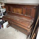 Hnědé dřevěné piano s výraznou masivní konstrukcí a panelovými výplněmi. Povrch nástroje je pokryt vrstvou prachu a vykazuje známky opotřebení. Na vi…