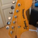 Záběr na hlavu kytary z přírodního dřeva s nápisem Squier JAGUAR by Fender. Na hlavě jsou umístěny šest kovových ladiček. Povrch dřeva je lakovaný a…
