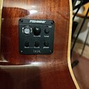 Detail předzesilovače s ladičkou zabudovaného do dřevěného těla akustické kytary. Černý ovládací panel nese nápisy FISHMAN, Volume, tuner, batt, bass…