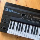 Černý syntetizátor s bílými a černými klávesami. Na horním panelu je nápis Roland D-50 LINEAR SYNTHESIZER. Jsou zde vidět různé ovládací tlačítka, po…