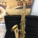 Zlatý saxofon umístěný v otevřeném černém kufru. Na mouthpiece je viditelný nápis V16. Povrch nástroje vykazuje známky opotřebení a drobné škrábance.