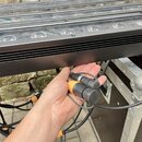 Detailní záběr na zadní část černého lineárního osvětlení s řadou LED diod. Ruka drží dva černé konektory, z nichž jeden má oranžový plastový prstene…