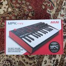 Fotografie zobrazuje krabici od MIDI kontroléru AKAI MPK mini. Na balení je zobrazen červený nástroj s klávesami a padem, doprovázený nápisy AKAI PRO…