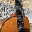 Detail akustické kytary s hnědým trupem a tmavým hmatníkem. Na povrchu desky jsou viditelné drobné škrábance a opotřebení laku. Na fotce nejsou žádná…