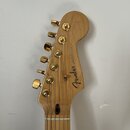 Hlava elektrické kytary z přírodního světlého dřeva se zlatými ladičkami. Na dřevě je vidět nápis Fender a vedle něj menší text STRATOCASTER. Povrch…