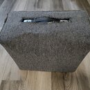 Šedý textilní kryt na hudební zesilovač nebo reproduktor. V horní části je výřez, ze kterého vykukuje černá plastová rukojeť. Okraje krytu jsou v něk…