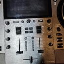 Detailní pohled na ovládací panel DJského kontroléru s kovovým povrchem a displejem. Na panelu jsou vidět tlačítka BACK, VIEW, BROWSE, LOAD a HOLD, p…