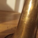 Detailní záběr na kovovou část hudebního nástroje v mosazném provedení. Povrch vykazuje známky opotřebení, škrábance a patinu. Na povrchu je vyryt ne…