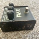 Černý kytarový efekt s nápisem RAT. Na horní straně jsou tři ovládací knoflíky s popisky VOLUME, FILTER a DISTORTION. Přístroj vykazuje výrazné opotř…