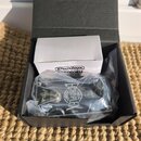 Černá papírová krabice, ve které se nachází bílá krabička s nápisem Dunlop ADB003EU ADAPTER. Vedle ní je v průhledném plastovém sáčku kytarový efekt…
