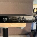 Černý audio interface s nápisem ensemble Thunderbolt™ Audio Interface. Zařízení má ovládací knoflík OUTPUT, čtyři tlačítka označená jako ASSIGNABLE (…