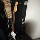 Elektrická kytara s tmavým tělem a bílým pickguardem, opřená o černý kufr. Na hlavě hředu je viditelné logo Fender a nápis STRATOCASTER. Kytara má hn…