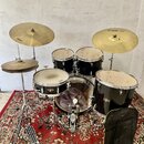Černá bubnová sada značky TAMA, která obsahuje basový buben, dva tom-tomy, malý tom a snare drum. Součástí jsou tři činely s nápisy PAISTE 202 a jede…