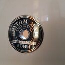 Detail na kulatý kovový štít s nápisy umístěný na bílém povrchu bubnu. Na štítu je kroužkově napsáno RHYTHM MATE a MADE IN CHINA, ve spodní části je…