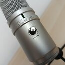 Detailní záběr na stříbrný kondenzátorový mikrofon s kovovou síťkou. Na těle je vidět nápis Superlux, zelená kontrolka a otočný knoflík s označením V…