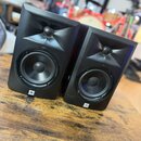 Dva černé studiové reproduktory umístěné na dřevěném stole. Na spodní části obou reproduktorů je bílé logo JBL. Povrch je matný a v horní části se na…