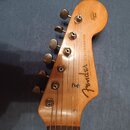 Záběr na dřevěnou hlavu elektrické kytary s šesti ladicími kolíky. Na povrchu je černý nápis Fender STRATOCASTER a menší text with synchronized tremo…