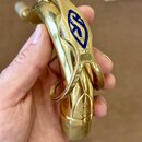 Detail zlatého saxofonu v ruky člověka. Na horní části je modré logo ve tvaru stylizovaného písmena S. Dole na zvonu je vyryté nečitelné logo a nápis…
