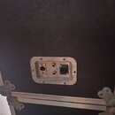 Detail zadní strany černého transportního kufru s texturovaným povrchem a hliníkovými hranami. Uprostřed je kovový panel s třemi konektory: jedním ko…