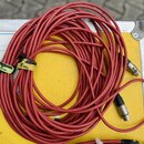 Červený kabel s kovovými konektory, svinutý do klubíčku na žlutém povrchu transportního kufru. Na kabelu jsou dvě žluté pásky s ručně psaným nápisem…