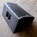 Černý stage monitor s kovovou mřížkou v přední části. Na pravém horním rohu mřížky je umístěno malé modré kulaté logo. Povrch je potažen černým textu…