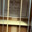 Fotografie detailu vnitřní štítku akustické kytary. Na štítku je logo SIGMA s nápisem EST.1970, model DM12E+ a sériové číslo 180106747. Dole je uvede…