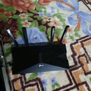 Na fotografii je černý plastový router se čtyřmi externími anténami. Na přední straně je viditelné bílé logo TP-Link. Zařízení leží na vzorovaném kob…