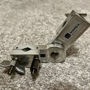Kovová svorka stříbrné barvy s nastavitel०m kloubem. Na boku je černý nápis MAPEX™ a pod ním menší text MADE IN CHINA. Povrch vykazuje drobné škrában…