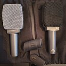 Dvě dynamická mikrofony s mřížkou, jeden v černé a druhý v stříbrné barvě. Na černém mikrofonu je nápis SENNHEISER MADE IN GERMANY a značka CE, na st…