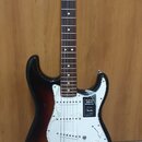 Elektrická kytara s tłem v barvě sunburst a bílým pickguardem. Na těle je nalepený štítek s nápisy PLAYER SERIES STRATOCASTER a logo Fender. Kytara m…