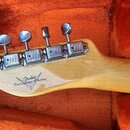 Zadní strana hlavy kytary z přírodního dřeva s šesti chromovanými ladičkami. Na dřevě je viditelný nápis Fender Custom Shop. Nástroj leží v otevřeném…