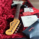 Elektrická kytara s tělem v provedení sunburst a světlou hmatníkem leží v otevřeném kufru s červeným plyšovým vnitřkem. Na hlavě kytary je viditelné…