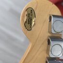 Detail zadní strany dřevěné hlavy kytary s kovovými ladičkami. Na dřevě je nalepený oválný zlatý štítek s nápisem MATT UMANOV GUITARS NEW YORK, který…