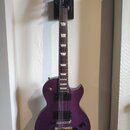 Fialová elektrická kytara s tvarem Les Paul, umístěná v nástěnním držáku. Na hlavě hředu je zlatý nápis Gibson. Kytara má dva humbuckery, čtyři ovlád…