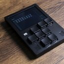 Černý elektronický sekvencer s displejem a mřížkou 16 gumových padů. Na spodní části šasi je bílý nápis „B R M B“. Displej zobrazuje textové menu s n…