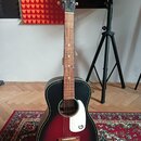 Akustická kytara s tělem v červeně-černém přechodu (sunburst) a bílým plechtrem, na kterém je černé logo „G“. Na hlavě hředu je bílý nápis „GRETSCH“.…