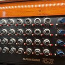 Černý mixovací pult s ovládacími knoflíky v mřížce 4x8 a modrými posuvníky. Na panelu jsou bílé nápisy s číslováním kanálů od 1 do 32 a označení skup…