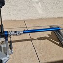 Kovový mechanismus pedálů na bicí soupravu v stříbrné a modré barvě. Soustava obsahuje dvě černá šlapadla s pěnovými kryty a propojnou hřídel v modré…