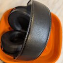 Černá sluchátka s polstrovaným náheadním pásem z materiálu imitující kůži. Sluchátka jsou umístěna v oranžovém plastovém obalu. Na viditelných částec…