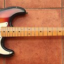 Elektrická kytara s tvarem Stratocaster, provedená v červeno-žlutém sunburst povrchu s bílým pickguardem. Na hlavě hředu je vidět logo Greco a nápis…