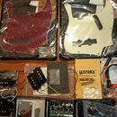 Sada příslušenství pro basovou kytaru rozložená na hnědém stole. V horní části jsou dvě pickguardy v plastových obalech značky FLEOR, jedna v červené…