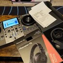 Na fotografii je černý DJ 컨트롤er značky Numark s modrým displejem a dvěma jogovými koly. Vedle něj leží krabice a černé sluchátka s nápisem Pioneer DJ…