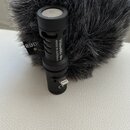 Černý mikrofon s nápisem VideoMic Me MADE IN AUSTRALIA. Je umístěn před šedým chlupatým windscreenem, na kterém je viditelné logo RØDE. V dolní části…