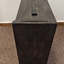 Tmavý dřevěný reproduktor nebo ozvučovací box s výraznou strukturou zrn. Na horní straně jsou umístěny dvě černé úchytky, jedna podélná a jedna popří…